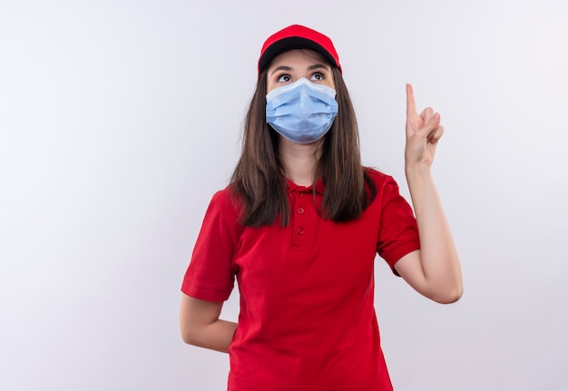 Young delivery girl wearing red t-shirt in red cap wears face mask points to up on isolated white background