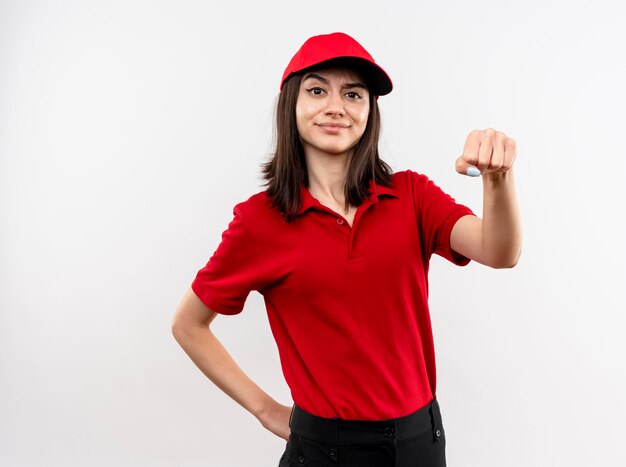 Young delivery girl wearing red uniform and cap clenching fist happy and positive smiling confident standing over white wall