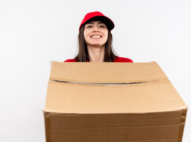 白い壁の上に立っている顔に笑顔で大きな段ボール箱を保持している赤い制服と帽子を身に着けている若い配達の女の子