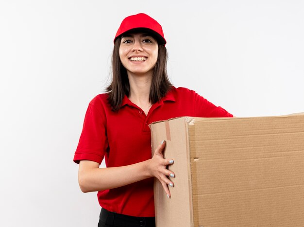 빨간 유니폼을 입고 젊은 배달 소녀와 흰 벽 위에 서있는 얼굴에 미소로 큰 골판지 상자를 들고 모자