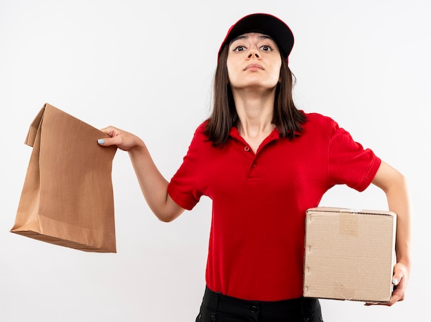 Young delivery girl wearing red uniform and cap holding paper package and cardboard box looking at camera with confident expression standing over white background
