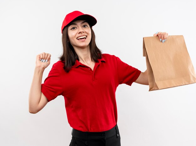Молодая доставщица в красной форме и кепке держит бумажный пакет, сжимая кулак, счастливая и взволнованная, стоя на белом фоне