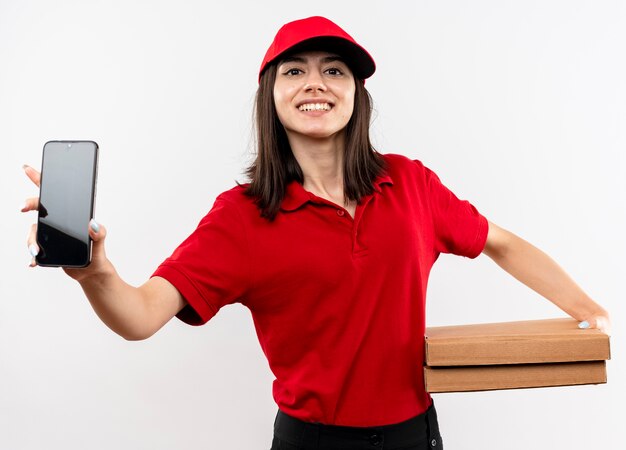 Молодая доставщица в красной форме и кепке держит коробки для пиццы, показывая смартфон, весело улыбаясь, глядя в камеру, стоящую на белом фоне