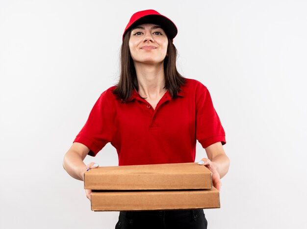 Young delivery girl wearing red uniform and cap holding pizza boxes smiling with happy face standing over white wall