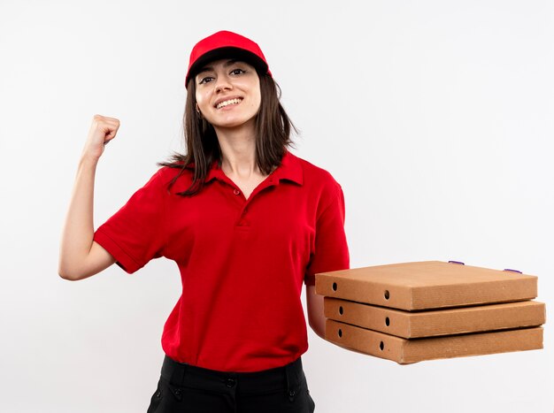 Young delivery girl wearing red uniform and cap holding stack of pizza boxes clenching fist happy and positive smiling standing over white background