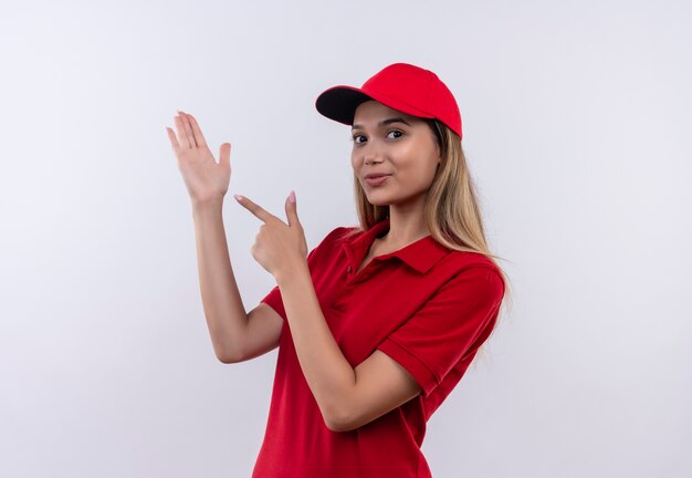 young delivery girl wearing red uniform and cap points to her hand  isolated on white wall