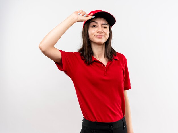 赤い制服とキャップを身に着けている若い配達の女の子は、白い壁の上に立っている彼女のキャップに触れて微笑んでウインク