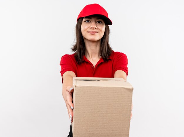 赤い制服と白い壁の上に立ってフレンドリーな笑顔のボックスパッケージを伸ばしてキャップを身に着けている若い配達の女の子