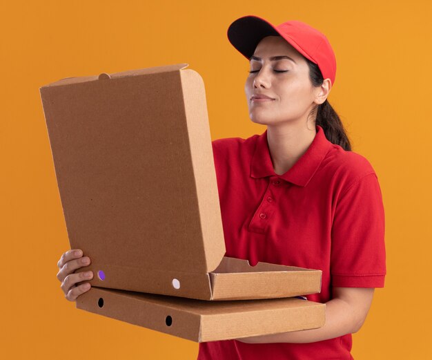 Young delivery girl wearing uniform and cap opening and sniffing pizza box isolated on orange wall