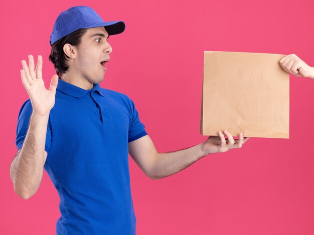 Young delivery man in blue uniform and cap isolated on pink wall