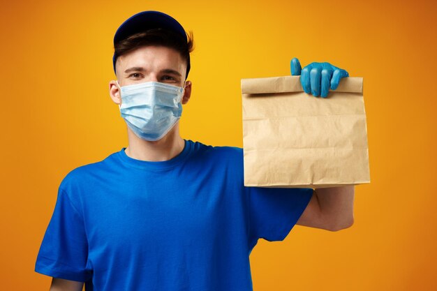 Young delivery man in face mask and gloves holding food delivery parcel against yellow background