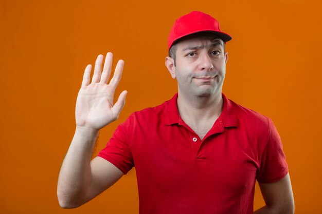 Бесплатное фото Молодой курьер в красной рубашке поло и кепке, стоящий с открытой рукой, делает знак остановки с серьезным и уверенным жестом защиты на изолированном оранжевом фоне на изолированном оранжевом