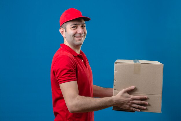 빨간 폴로 셔츠와 모자 고립 된 파란색 벽 위에 친절 한 미소 고객에 게주는 골 판지 상자를 들고 젊은 배달 남자