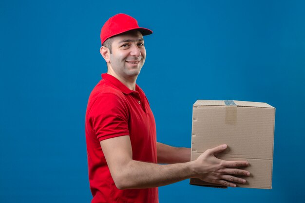 Young delivery man in red polo shirt and cap holding cardboard box giving to a customer smiling friendly over isolated blue wall