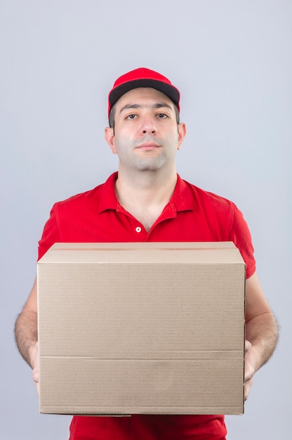 Молодой доставщик в красной рубашке поло и кепке держит картонную коробку с серьезным лицом, стоя над изолированной белой стене