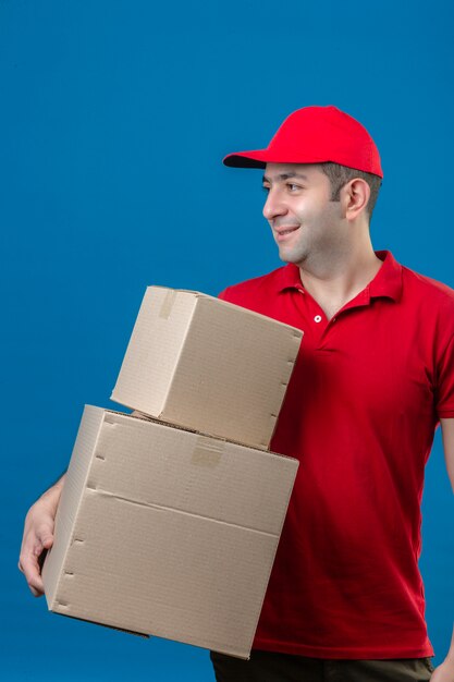 빨간 폴로 셔츠와 모자 고립 된 파란색 벽 위에 서 얼굴에 미소 옆으로 찾고 골판지 상자를 들고 젊은 배달 남자