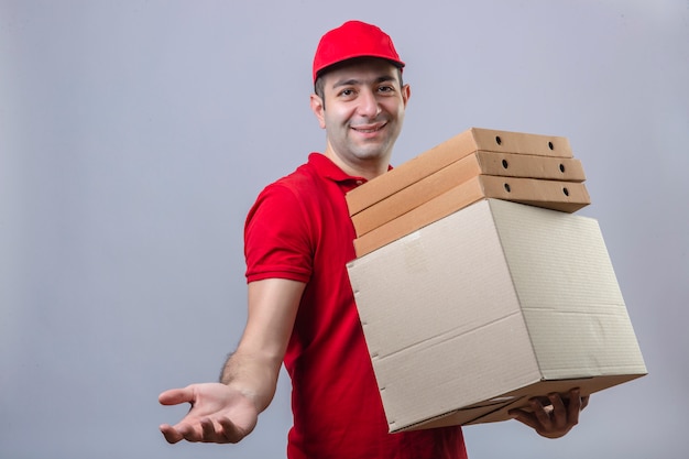 Young delivery man in red polo shirt and cap holding cardboard boxes smiling making gesture with hand waiting for a payment over isolated white wall