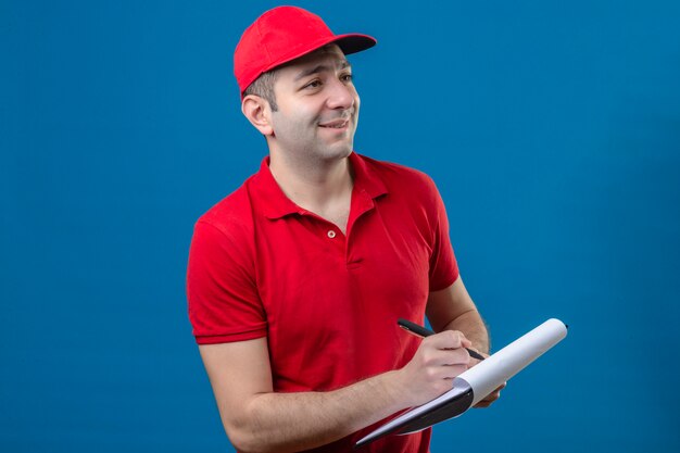 Молодой доставщик в красной рубашке поло и кепке держит буфер обмена, писать что-то смотрит в сторону с улыбкой на лице над синей стеной