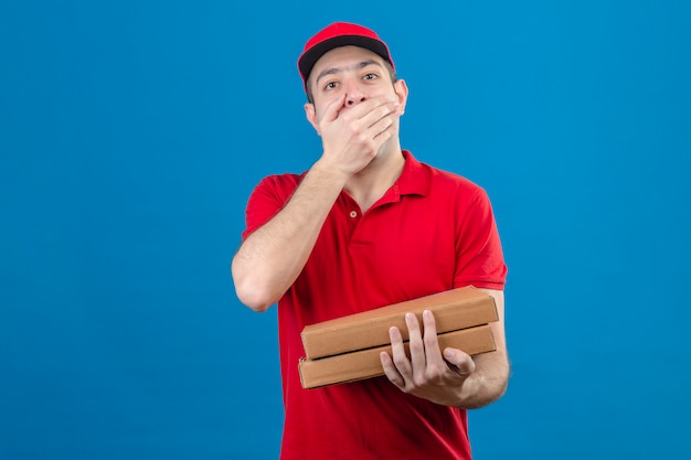 Молодой доставщик в красной рубашке поло и кепке держит коробки для пиццы, прикрывая рот рукой в шоке