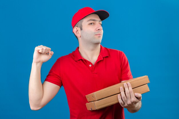 Young delivery man in red polo shirt and cap holding pizza boxes looking confident raising fist after a victory winner concept standing over isolated blue wall