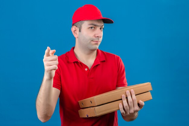 Young delivery man in red polo shirt and cap holding pizza boxes looking reproachfully at the camera and pointing a finger at you over isolated blue wall