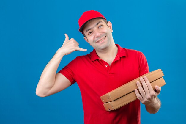 빨간 폴로 셔츠와 모자 피자 상자를 들고 젊은 배달 남자 전화 나 고립 된 파란색 벽 위에 친절 한 미소 전화