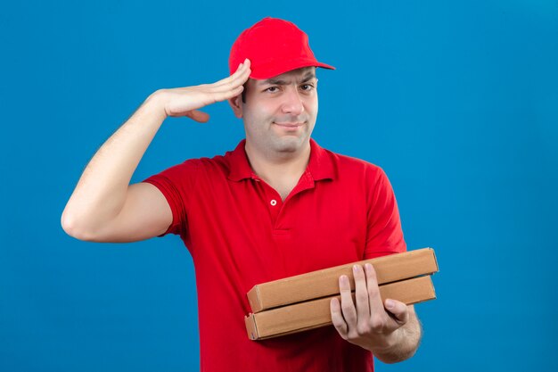 赤いポロシャツと分離のオレンジ色の壁に深刻な顔で立っている仕事の準備ができて敬礼を作るピザの箱を保持しているキャップの若い配達人
