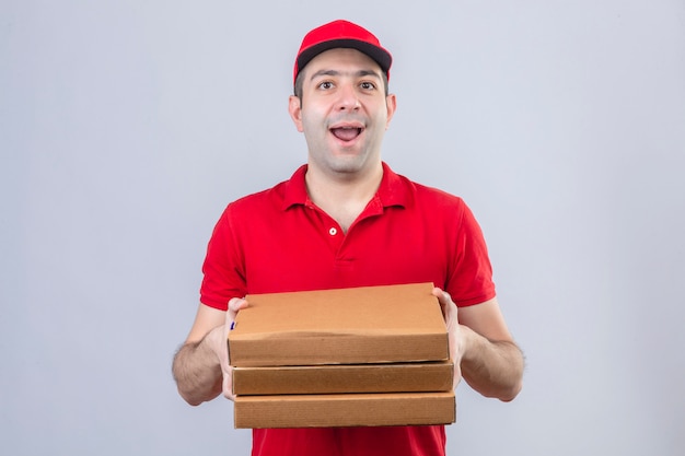 Young delivery man in red polo shirt and cap holding pizza boxes smiling cheerfully standing over isolated white wall