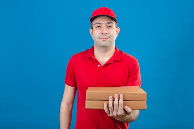 Young delivery man in red polo shirt and cap holding pizza boxes smiling friendly standing over isolated blue wall