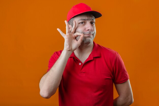 빨간색 폴로 셔츠와 모자에 젊은 배달 남자 격리 된 오렌지 배경 위에 지퍼로 입을 닫는 것처럼 침묵 제스처를 만드는