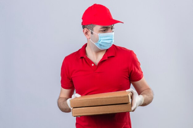 赤いポロシャツの若い配達人と分離の白い壁を恐れて恐れてよそ見ピザの箱を保持している医療マスクのキャップ