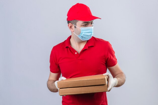 Young delivery man in red polo shirt and cap in medical mask holding pizza boxes looking aside with with fear over isolated white wall