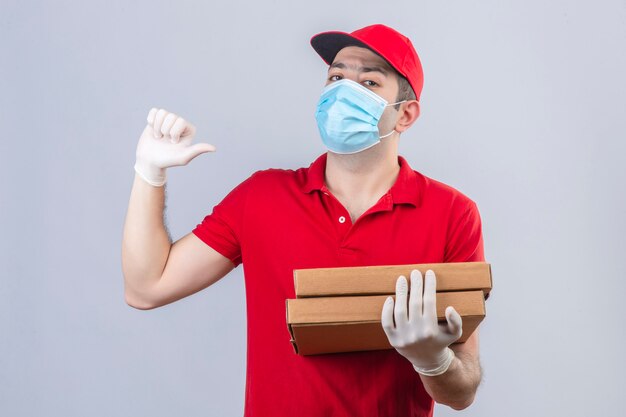 Молодой доставщик в красной рубашке поло и кепке в медицинской маске держит коробки для пиццы, глядя уверенно указывая на себя над изолированной белой стеной