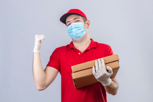 赤いポロシャツの若い配達人と手と親指で分離された白い壁に自信を持って見て後ろで後ろを指しているピザの箱を保持している医療マスクのキャップ