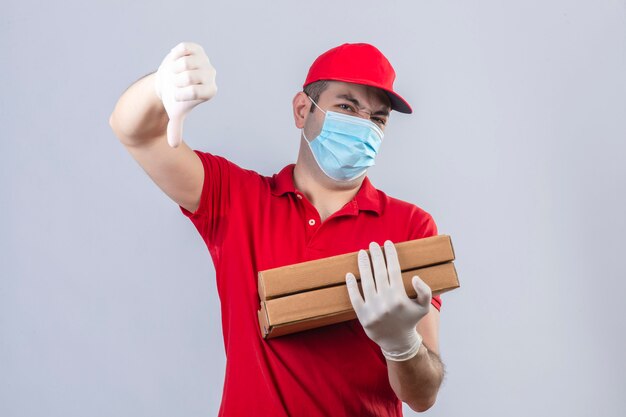 Young delivery man in red polo shirt and cap in medical mask holding pizza boxes standing with thumb down showing dislike over isolated white wall