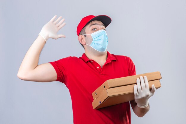 Young delivery man in red polo shirt and cap in medical mask holding pizza boxes waving with hand looking surprised over isolated white wall