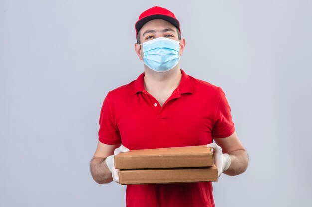 Young delivery man in red polo shirt and cap in medical mask holding pizza boxes with smile on face over isolated white wall