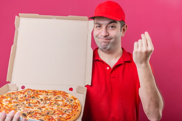 赤いポロシャツとキャップ立って分離のピンクの背景の上のカメラで手に笑顔でおいしいジェスチャーを作る新鮮なピザの箱で立っている若い配達人
