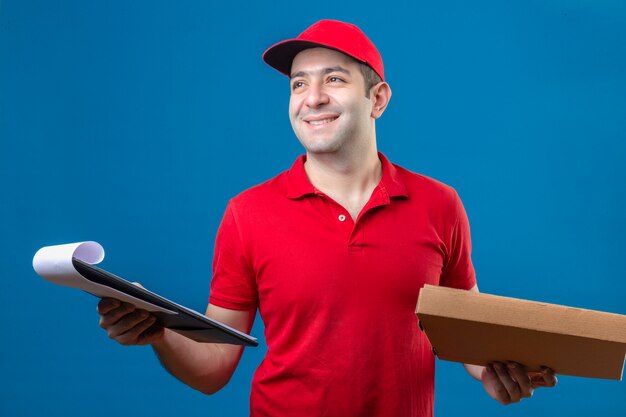 赤いポロシャツとピザの箱と孤立した青い背景に幸せそうな顔で優しい笑顔の手でクリップボードでキャップ立っている若い配達人