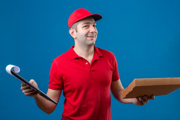 Young delivery man in red polo shirt and cap standing with pizza box and clipboard in hands smiling friendly with happy face over isolated blue background