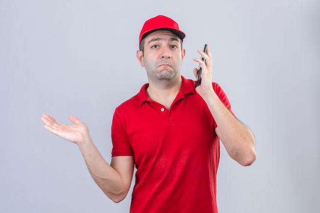 Young delivery man in red polo shirt and cap talking on mobile phone clueless and confused expression with arm raised having doubts over isolated white background