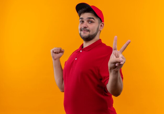 Young delivery man in red uniform and cap clenching fist showing victory sign smiling