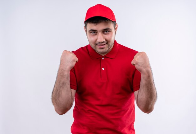 Young delivery man in red uniform and cap clenching fists happy and excited standing over white wall