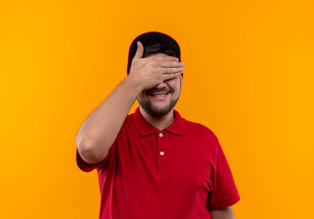 Young delivery man in red uniform and cap covering eyes with hand smiling waiting for surprise