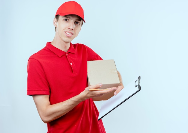 Young delivery man in red uniform and cap holding box package and clipboard looking to the front smiling confident standing over white wall