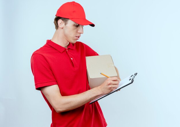 빨간 제복을 입은 젊은 배달 남자와 흰 벽 위에 서있는 심각한 얼굴로 뭔가 쓰는 상자 패키지와 클립 보드를 들고 모자