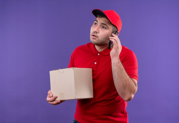 Young delivery man in red uniform and cap holding box package looking aside with serious face while talking on mobile phone standing over purple wall