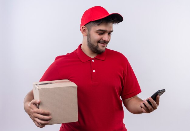 Young delivery man in red uniform and cap holding box package looking at screen of his smartphone smiling 