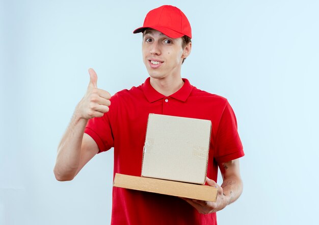 Young delivery man in red uniform and cap holding box package and pizza box looking to the front smiling showing thumbs up standing over white wall