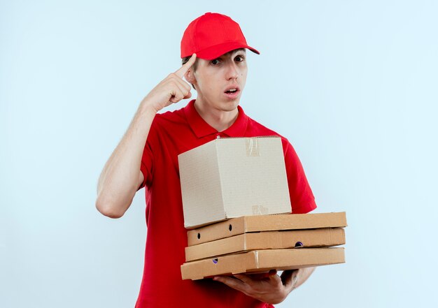 赤い制服と帽子を持った箱のパッケージとピザの箱を持った若い配達人が、白い壁の上に立っている仕事に集中して自信を持って彼の寺院を指しています